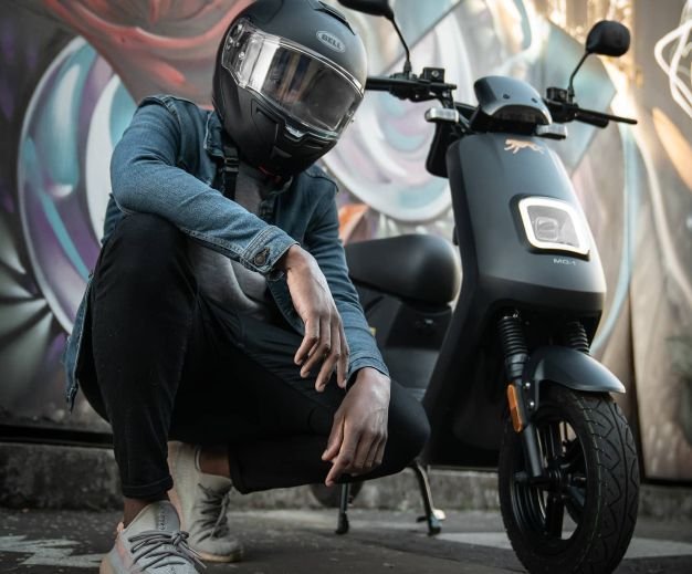 young hipster with his helmet possing alongside his Bilis electric moped. graffiti behind him.