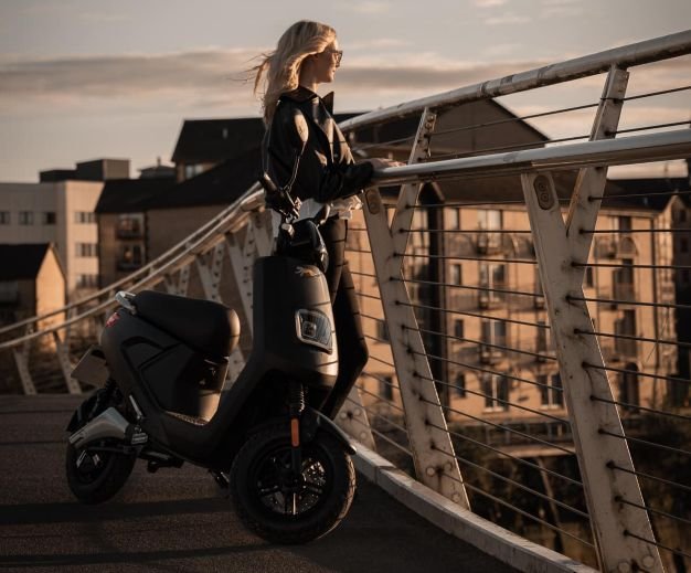 Young woman admiaring the sunset on a bridge alongside her Bilis electric moped